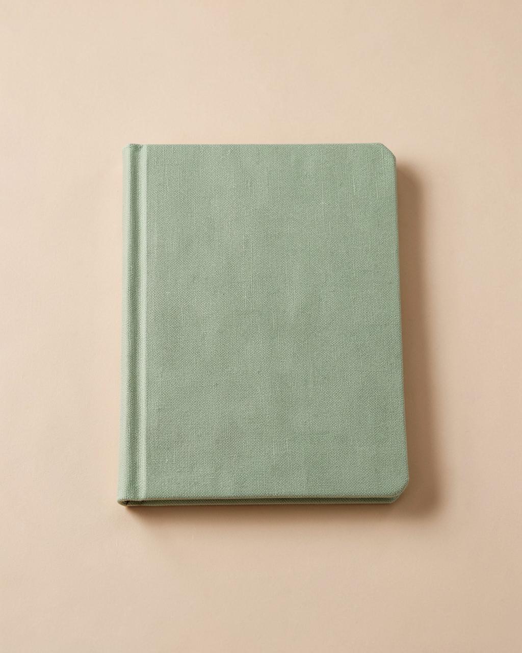 Carnet de routine matinale à couverture textile vert sauge posé sur fond beige clair